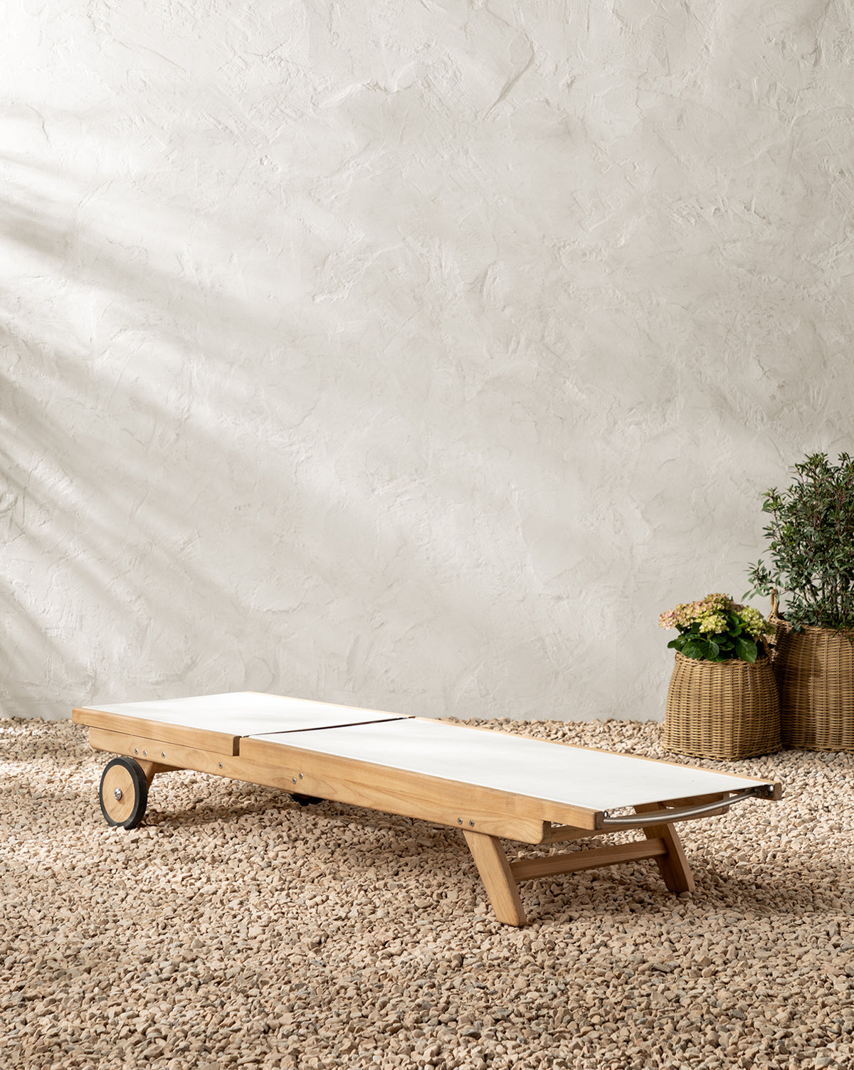 The Cadie Outdoor Chaise features white weather-resistant cushions and sits on gravel before a textured beige wall. Two woven baskets with green plants appear in the background on the right, by McGee & Co.