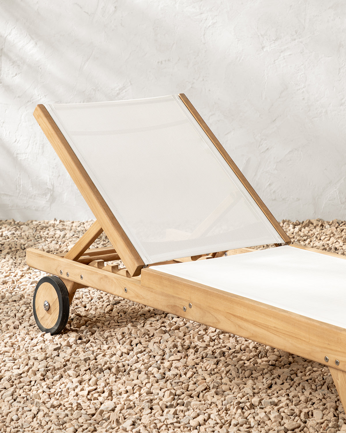 The Cadie Outdoor Chaise, featuring weather-resistant fabric on wood, rests on light gravel beside a textured white wall by McGee & Co.