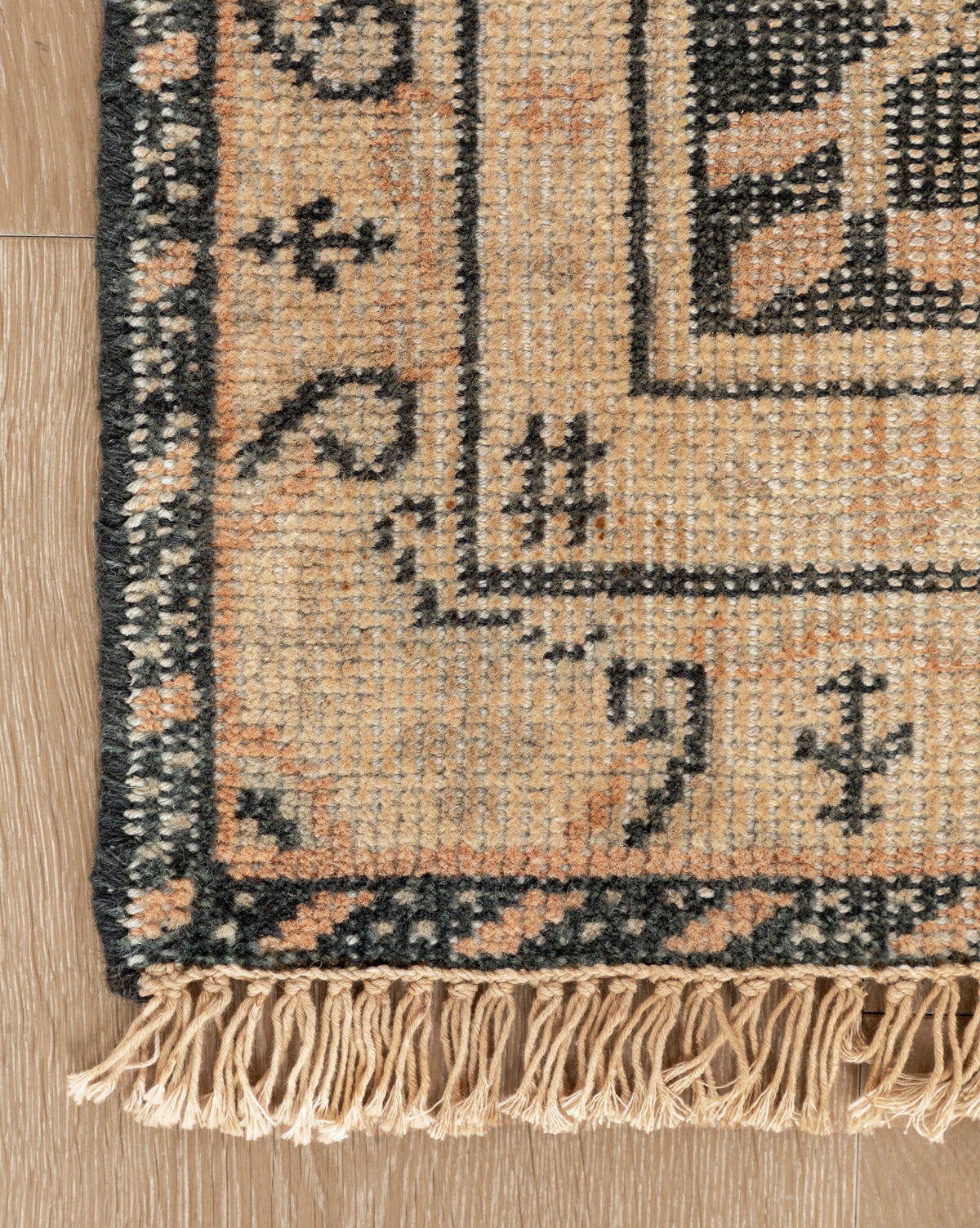 Close-up of the Campanella Hand-Knotted Wool Rug, showcasing a geometric border in beige, black, and peach tones with artisan fringe details on the edge, displayed on a wooden floor, by McGee & Co.