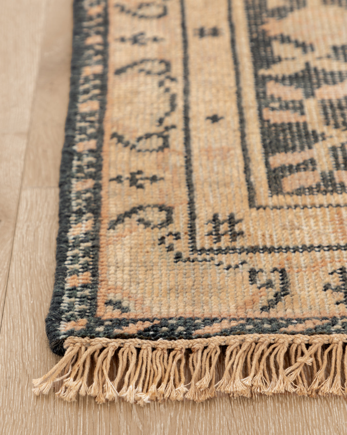 Close-up of the Campanella Hand-Knotted Wool Rug, showcasing intricate geometric patterns in beige and dark gray, artisan details, and edge fringe on a light wooden floor, by McGee & Co.