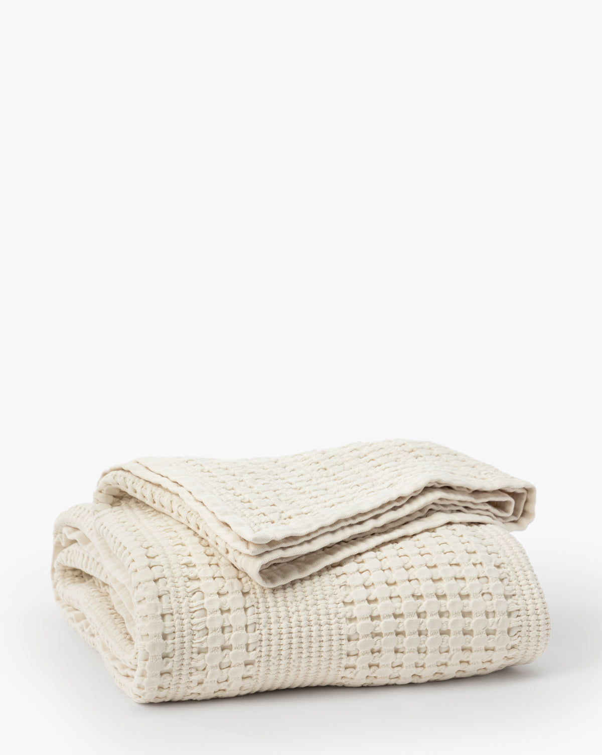 The Carnel Waffle Blanket, folded neatly against a plain white background, features a cozy textured pattern. Crafted in Portugal from 100% cotton, its cream color and inviting design add warmth to any space.