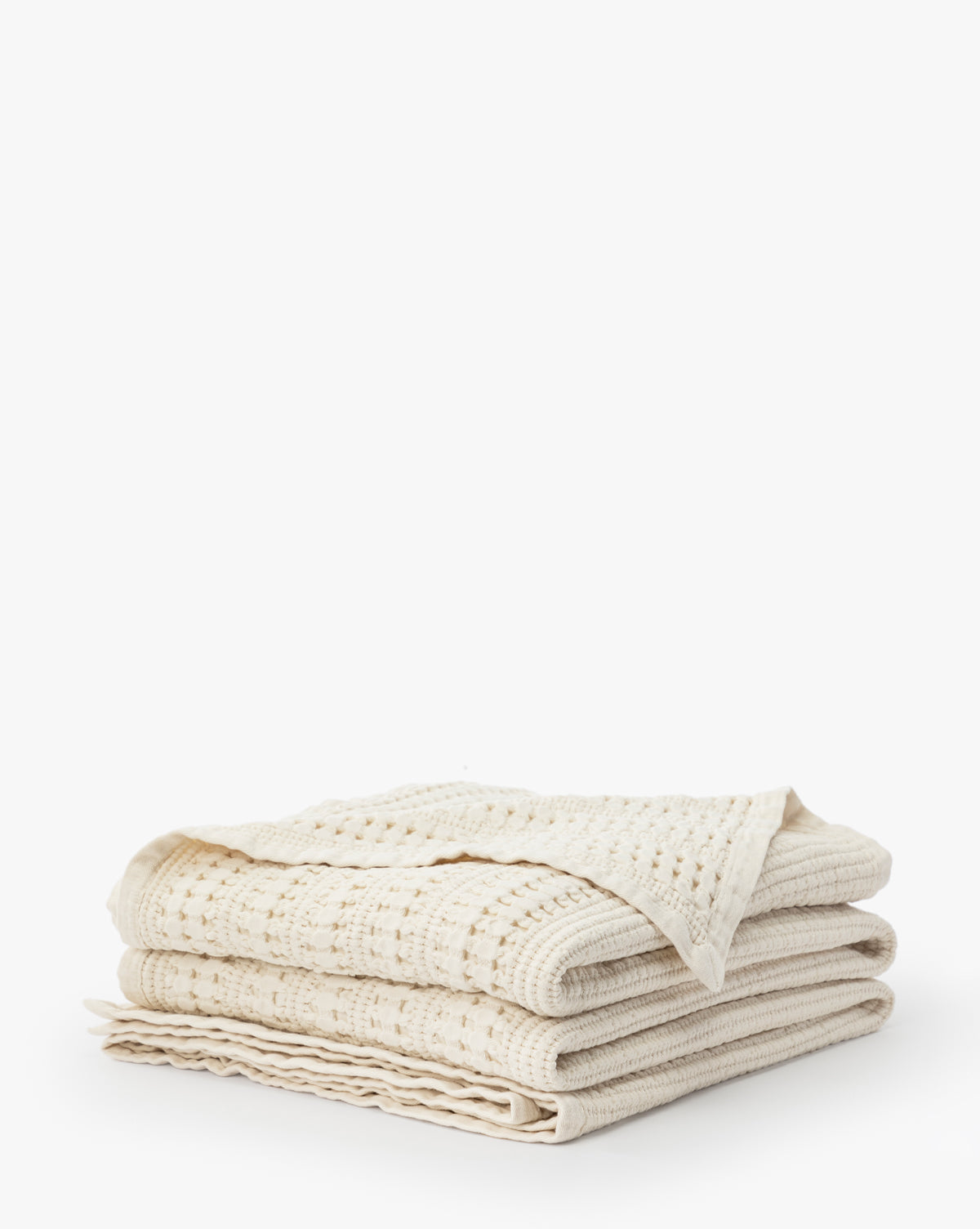The Carnel Waffle Blanket, made from 100% cotton and featuring a textured cream-colored design, is neatly folded and stacked on a white surface against a plain white background.