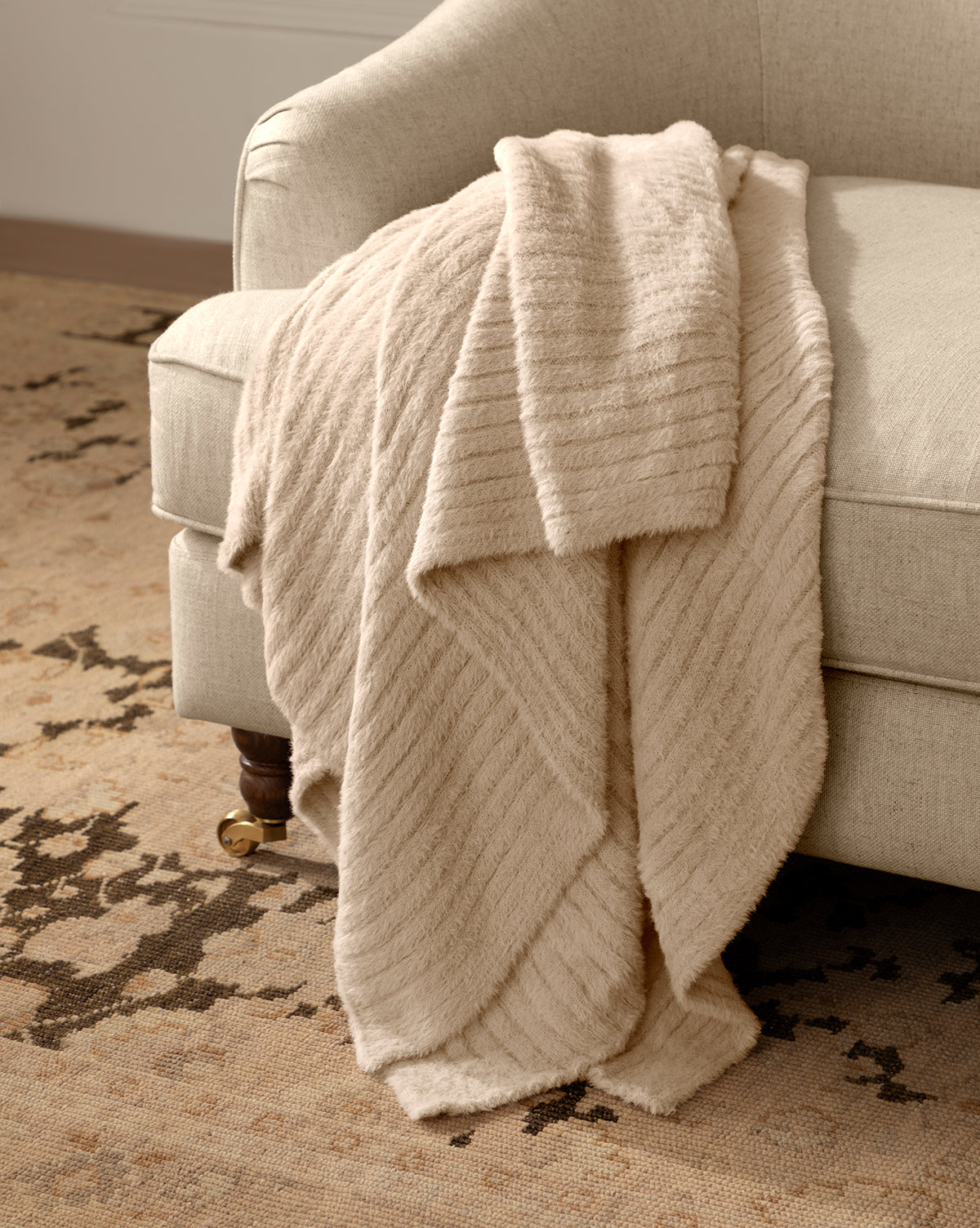 The soft, cream-colored Caro Throw is draped over the arm of a light beige upholstered chair, with a patterned beige and brown area rug underneath, by McGee & Co.