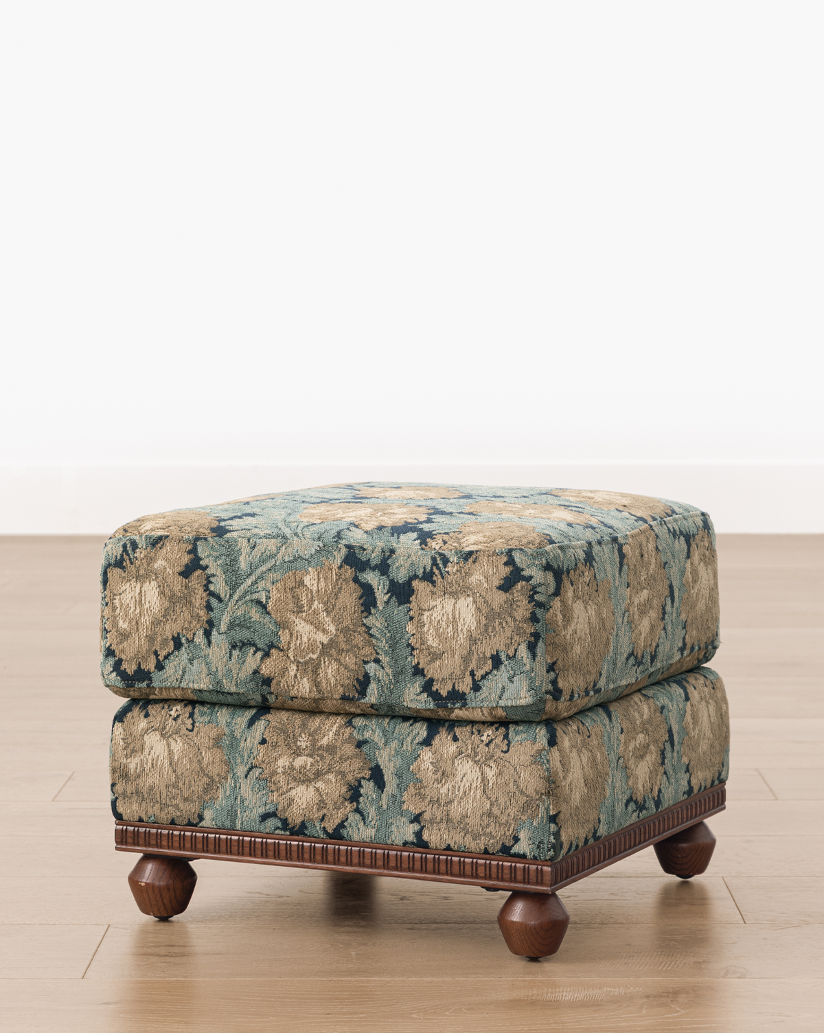 The Catherine Ottoman features a square, upholstered design with blue and beige floral fabric, a carved wood base, and round wooden feet, set on a light wood floor against a white background, by McGee & Co.