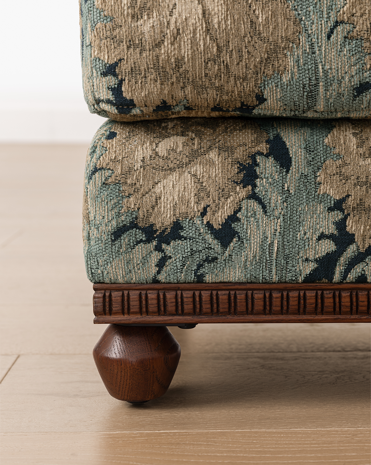 Close-up of the Catherine Ottoman, upholstered in green and beige patterned fabric, with a carved wood base and a round dark wooden leg, shown on a light wood floor, by McGee & Co.