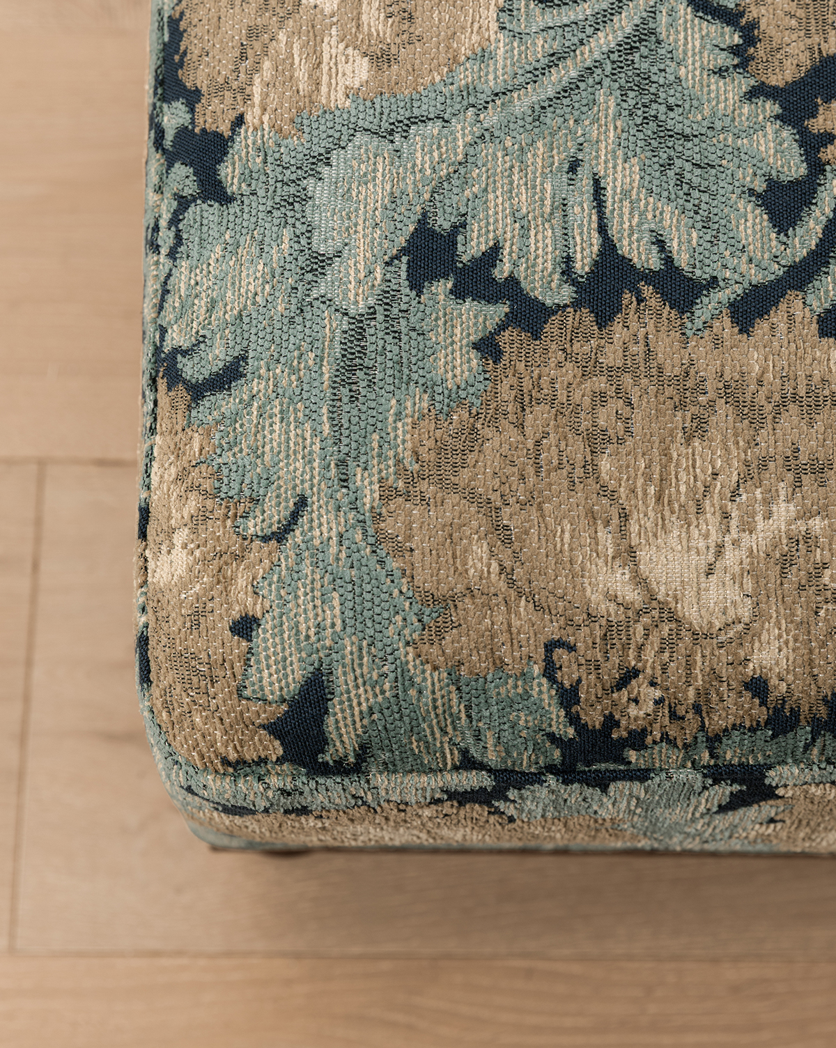 Close-up of the Catherine Ottoman corner, featuring green and gold floral patterned fabric with a carved wood base, on a light wooden floor, by McGee & Co.
