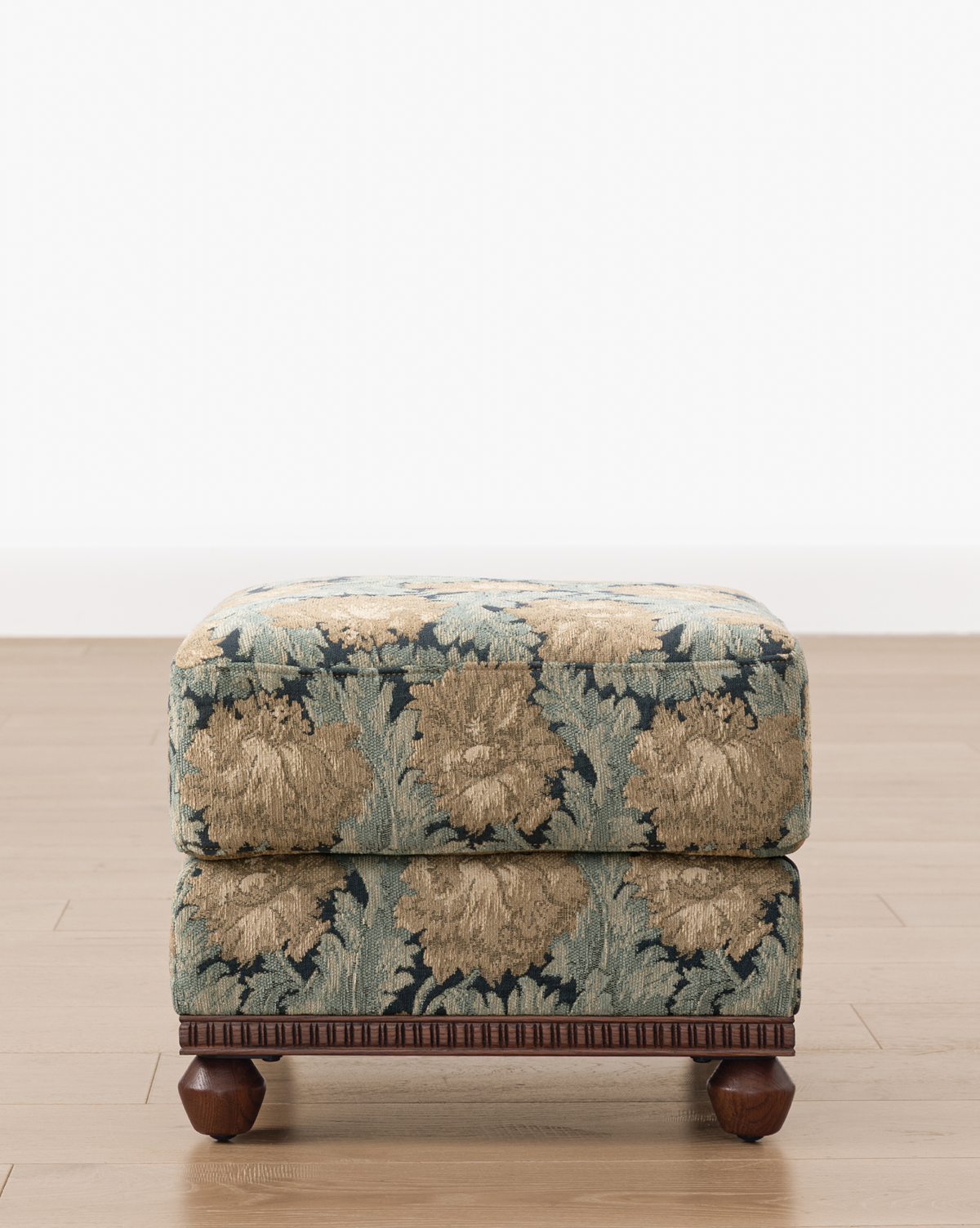 The Catherine Ottoman, featuring a carved wood base and ornate blue and beige floral upholstery, is placed on a light wood floor against a plain white wall, by McGee & Co.
