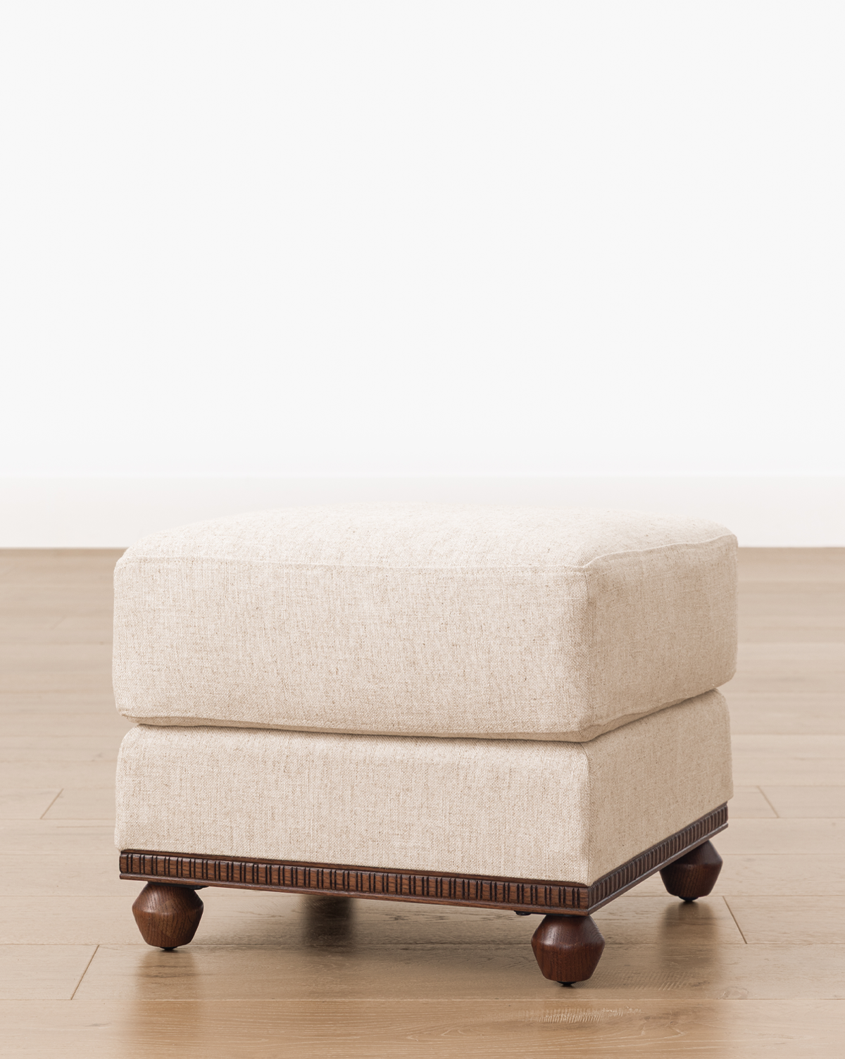 The Catherine Ottoman has a square beige upholstered top and dark carved wood base with four round wooden legs, adding elegance to any space, by McGee & Co.