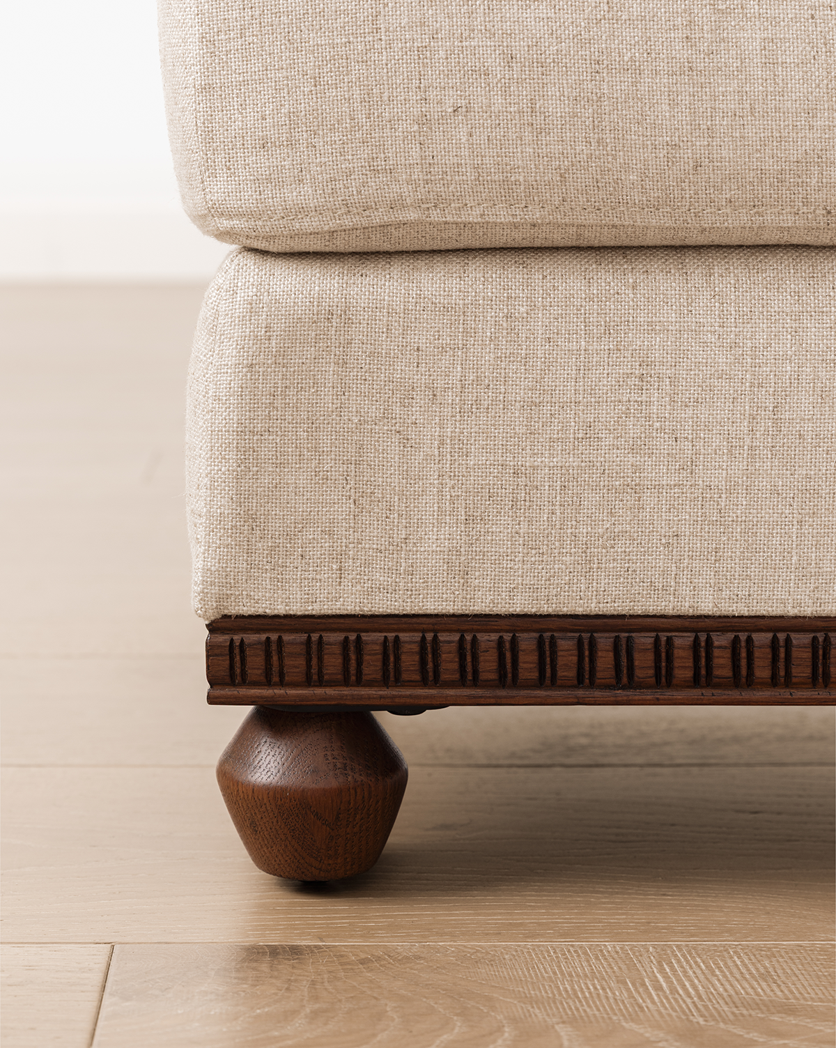 Close-up of a beige upholstered sofa with a carved wood base and round wooden leg, placed on a light wood floor beside the Catherine Ottoman, by McGee & Co.