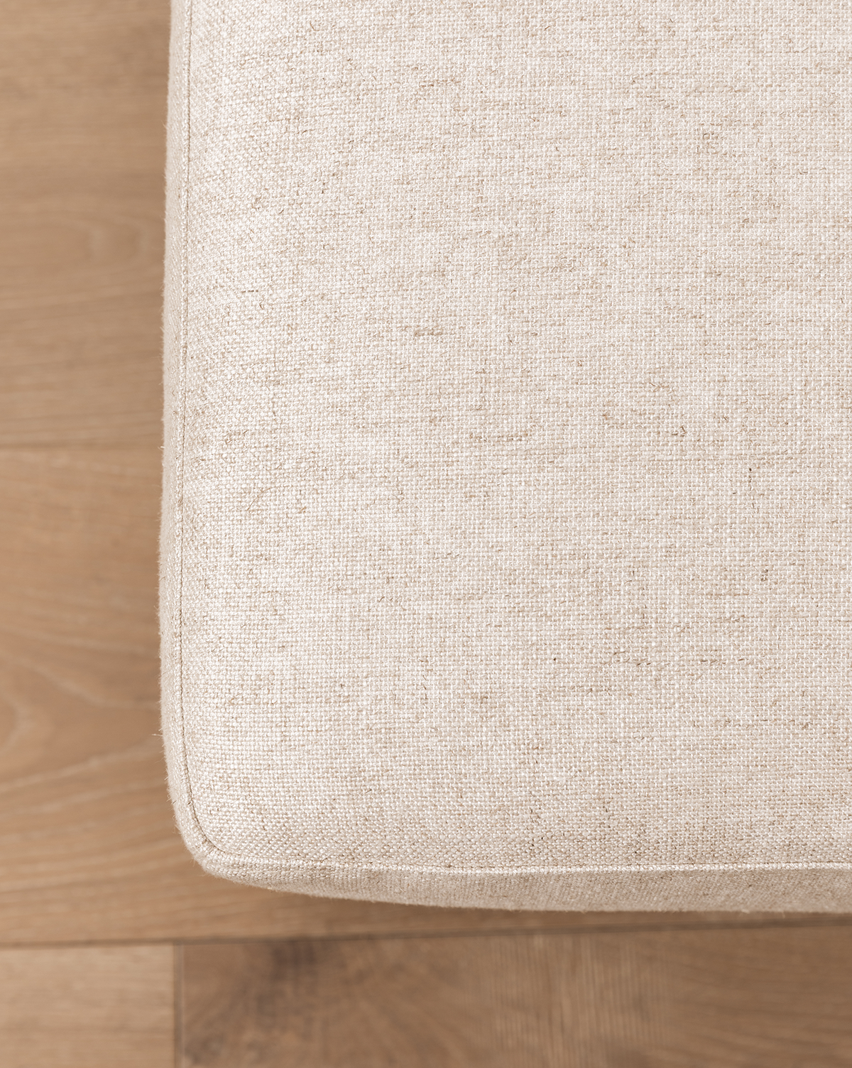 Close-up of the corner of a beige fabric cushion on the Catherine Ottoman, set on a light wooden floor. The upholstery texture and wood grain are clearly visible, by McGee & Co.