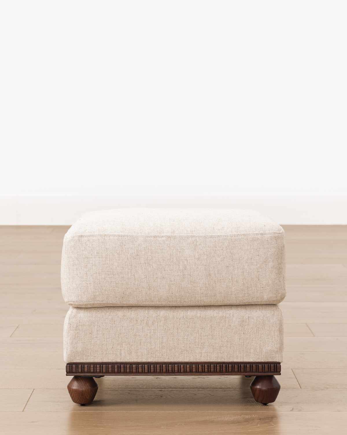 This upholstered ottoman looks stylish on a light wood floor beside a plain white wall, by McGee & Co.