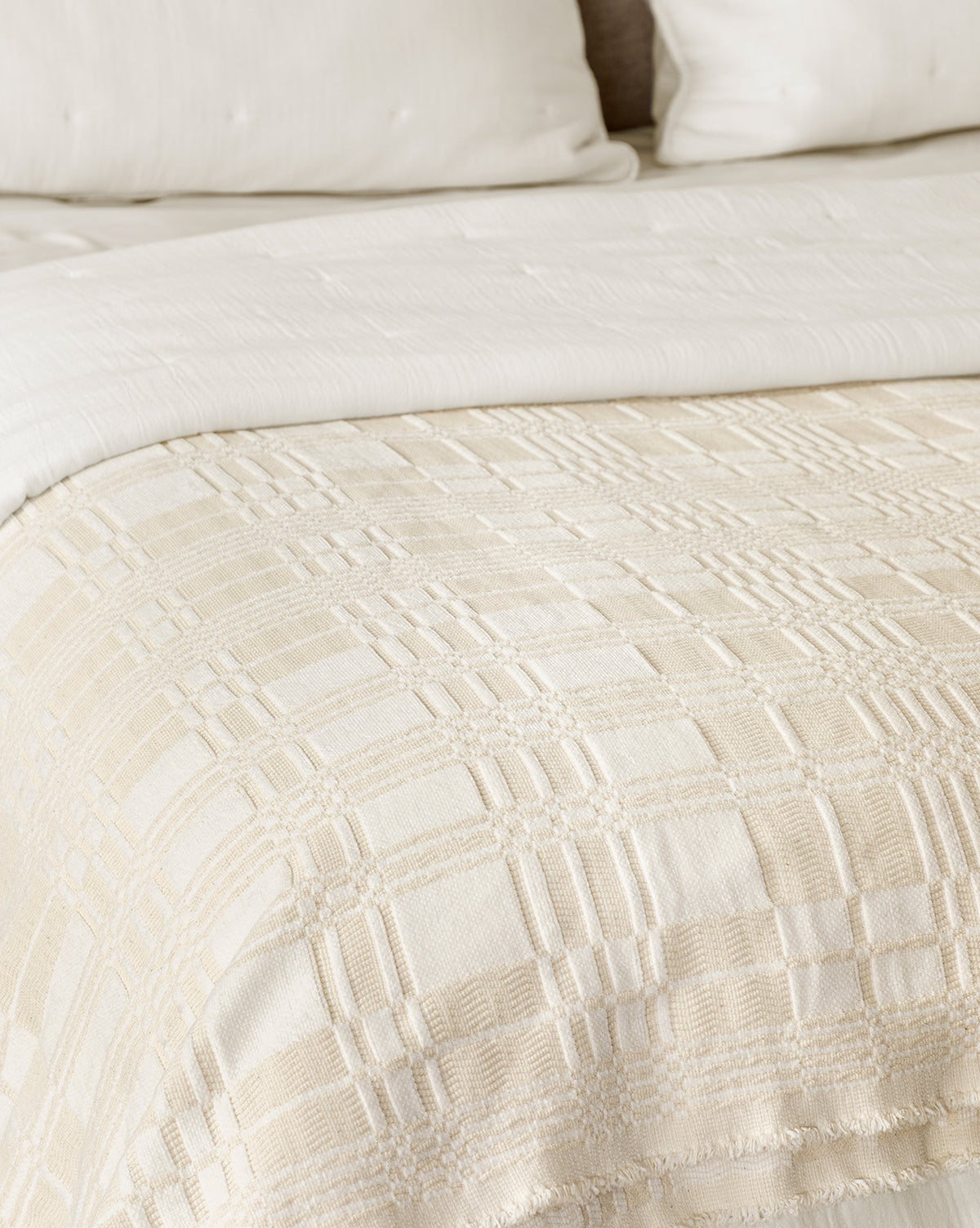 A close-up of a bed with cream bedding, showcasing the Makerspalm Chamberlain Coverlet, a textured cotton layer paired with a plaid woven blanket and matching pillows in the background, from McGee & Co.