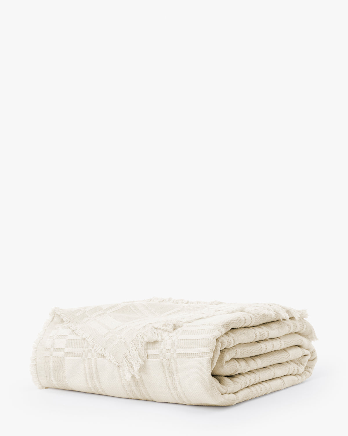 The Makerspalm Chamberlain Coverlet is a cream-colored woven blanket featuring a subtle checkered pattern and fringed edges, shown folded against a plain white background. McGee & Co.