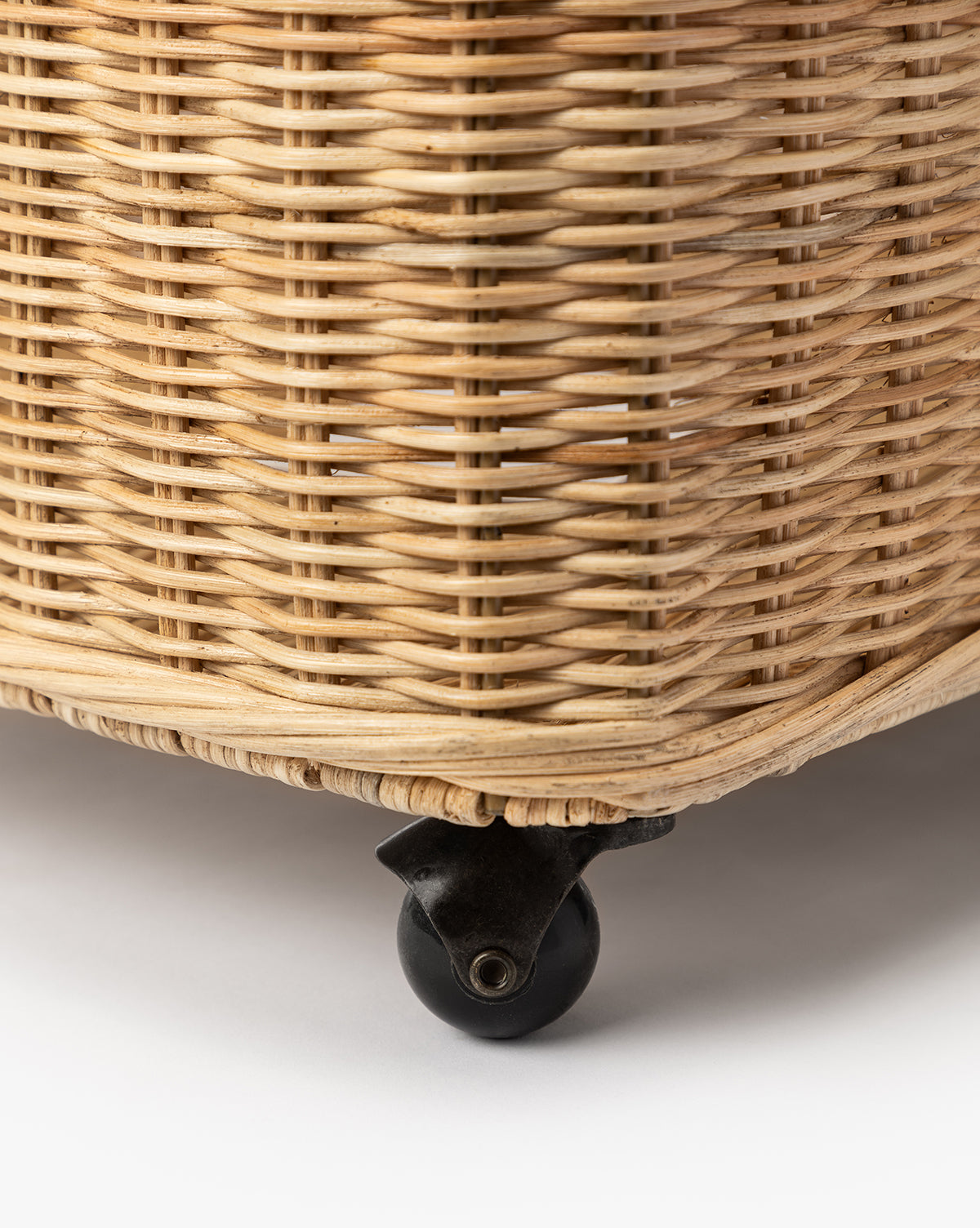 Close-up of the Chiara Laundry Basket in a natural wicker finish, featuring a black caster wheel attached to its bottom, placed on a white surface, by McGee & Co.