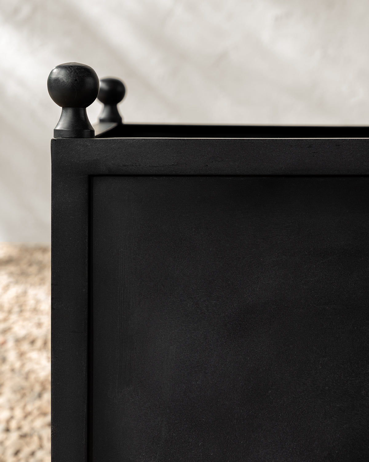Close-up of the Clariance Planter, a black rectangular metal design with rounded finials, set against a textured white wall and pebbled surface—ideal for refined outdoor styling.