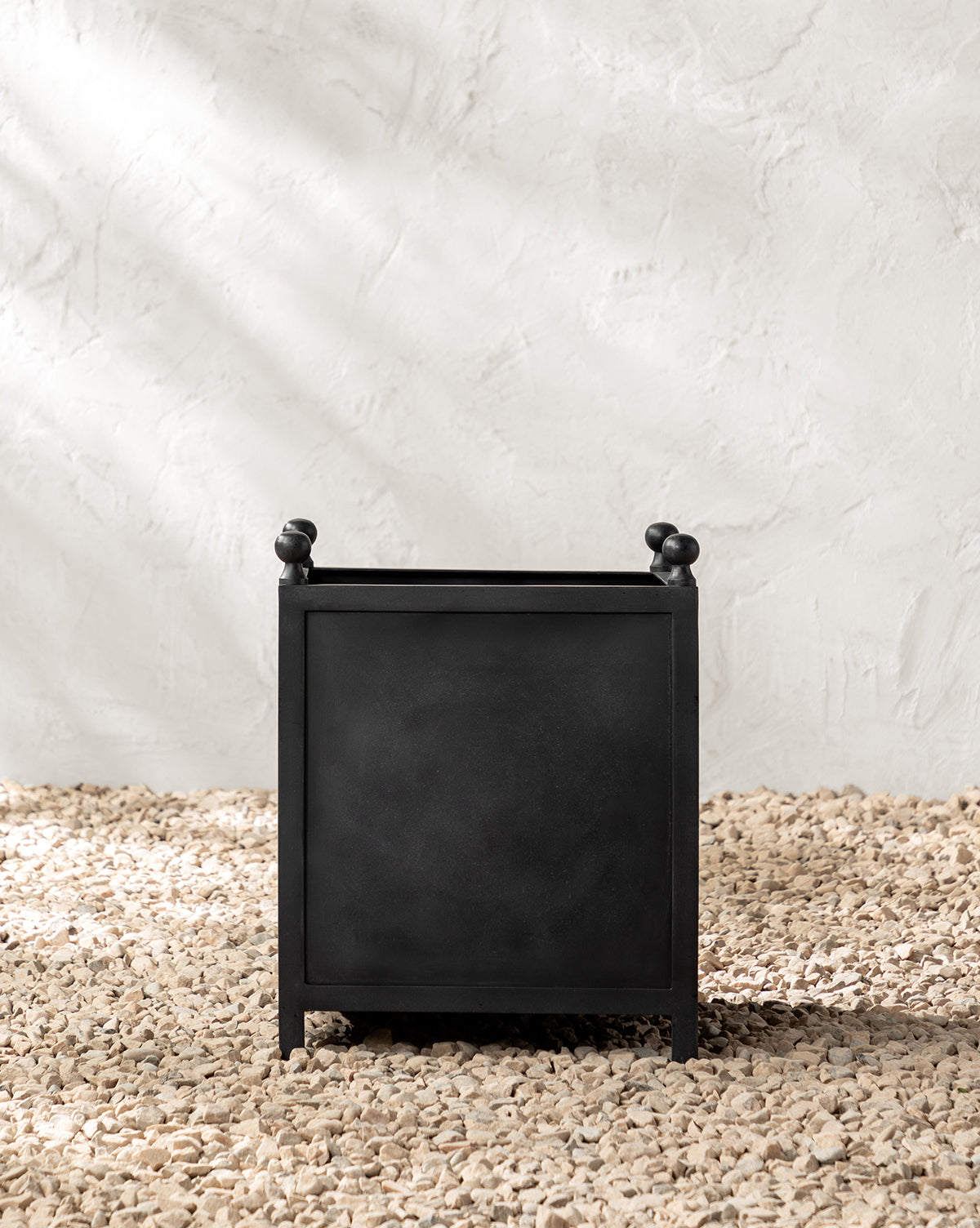 The Clariance Planter, featuring a square shape with small rounded corner knobs, rests on light pebbles, making it ideal for outdoor decor against a textured white wall.