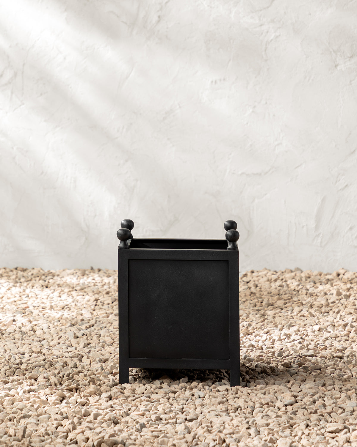 A black Clariance Planter with small rounded feet and decorative knobs rests on light pebbles against a textured white wall, bathed in sunlight—perfect for outdoor decor.
