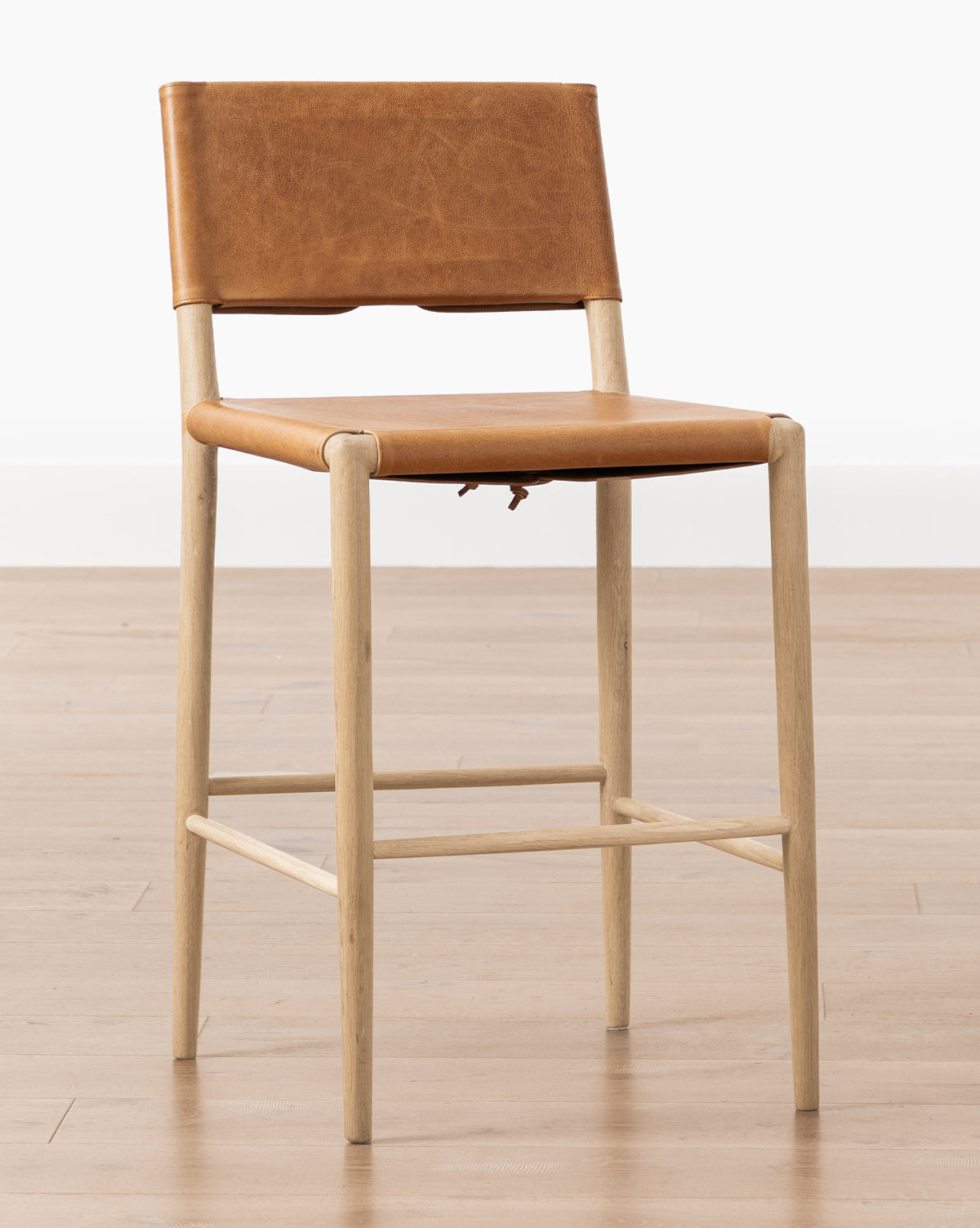 The Danson Counter Stool has a minimalist design with a light wooden frame, tan leather seat and backrest, shown on a light wood floor with a plain white background by McGee & Co.