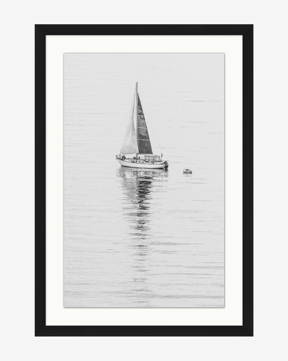 Early Voyage is a black and white photo featuring a sailboat with a single sail gliding on calm water, its reflection visible, with a small dinghy nearby. The striking image is elegantly framed in a simple black wood frame, by McGee & Co.