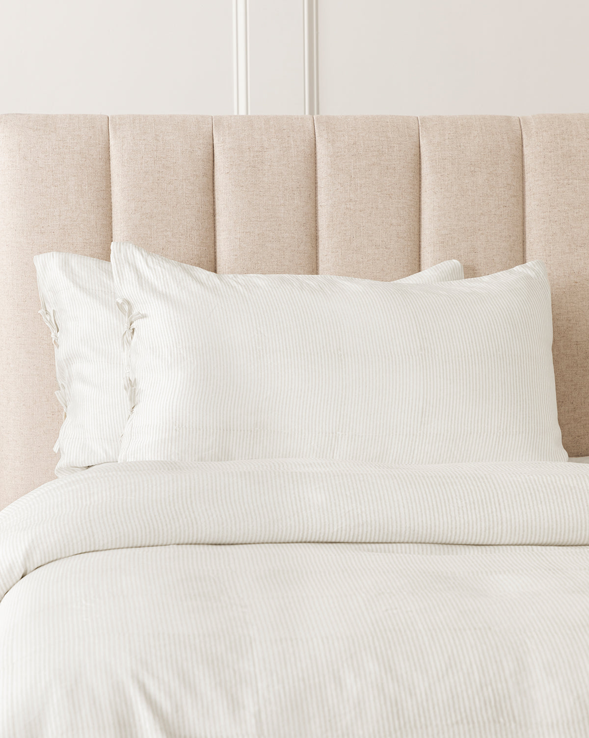 A neatly made bed with a light beige padded headboard, styled with Easton Striped Shams (Set of 2) and coordinating cream and oatmeal bedding, all in a softly lit bedroom for a timeless, classic look.