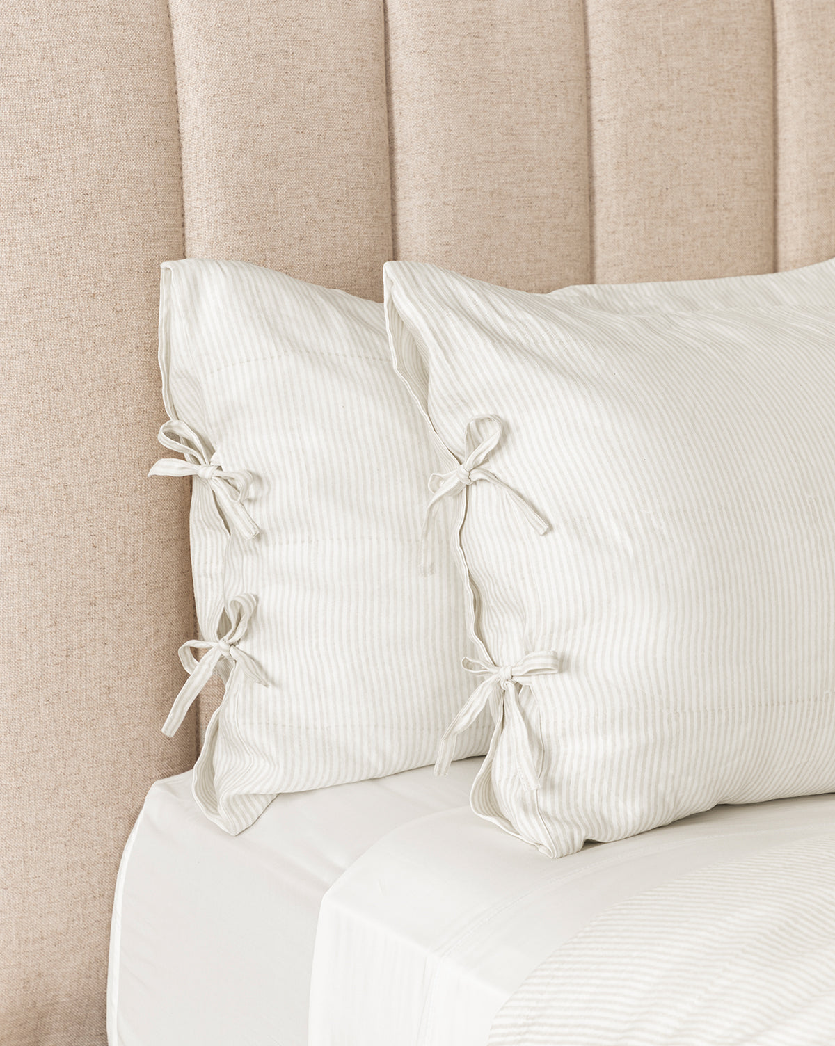 Two Easton Striped Shams (Set of 2) with subtle pinstripes and tied bow closures rest against a beige upholstered headboard, complementing classic design shams and cream and oatmeal bedding on a bed made with crisp white sheets.