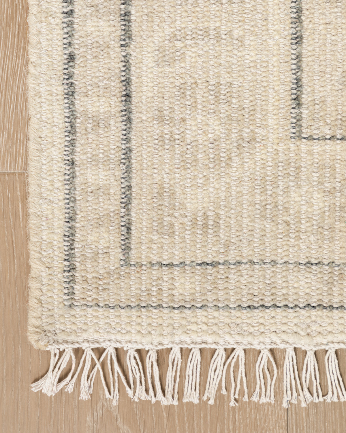 A close-up of the Elin Hand-Knotted Wool Rug highlights its simple gray border, artisan-crafted wool texture, and fringe details, beautifully displayed on a light wooden floor, by McGee & Co.