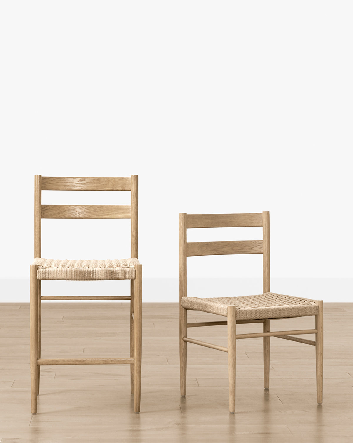 Two Eloise Woven Chairs, inspired by Danish design, are placed side by side on a light wooden floor against a white wall. The right chair is lower and smaller, adding stylish contrast to your dining room setting, by McGee & Co.