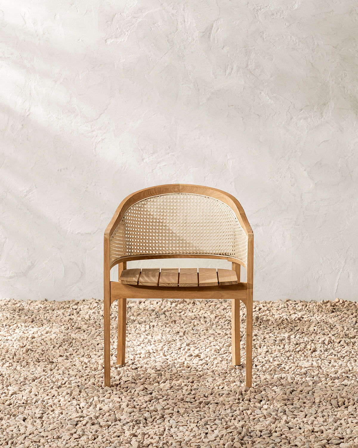 The Elowyn Outdoor Dining Chair, featuring a teak frame and woven cane backrest, sits on a textured pebble floor against a white textured wall in a minimalistic setting by McGee & Co.