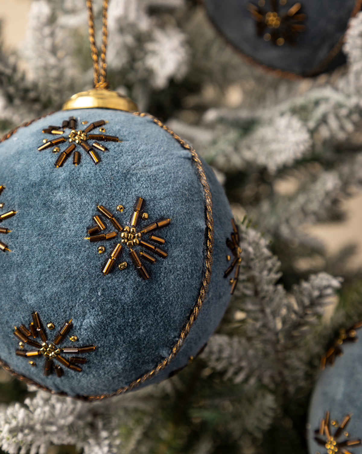 A 4" Embroidered Velvet Bauble from a set of 3, featuring blue velvet with gold beaded snowflakes, brings vintage elegance to a snow-dusted Christmas tree, by McGee & Co.