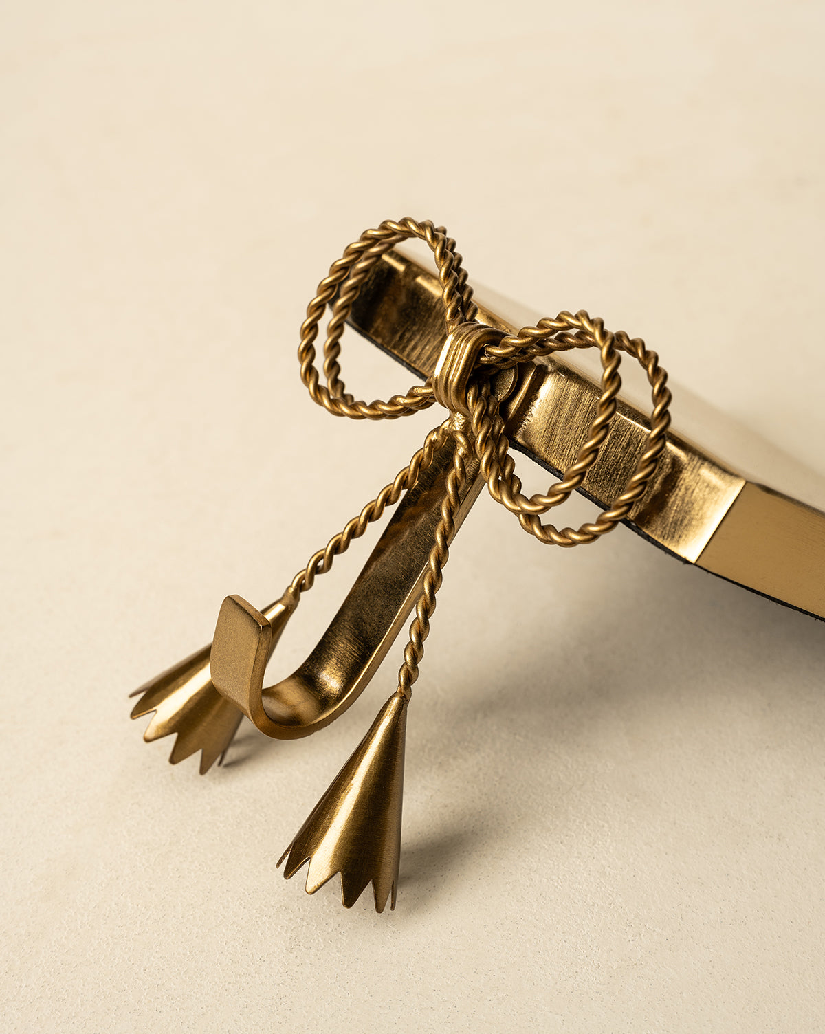 A gold metal book hook featuring a twisted rope bow and tassel design, inspired by the Erika Stocking Holder, rests on the corner of a closed book against a neutral background, by McGee & Co.