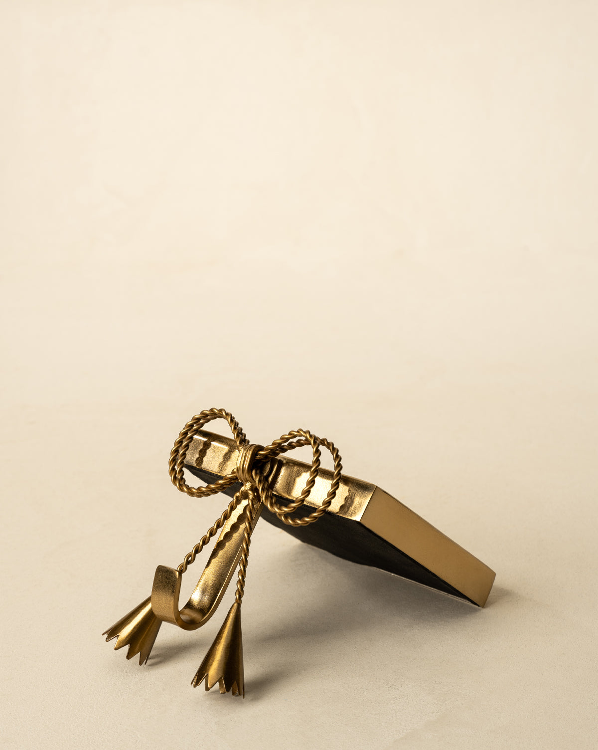The Erika Stocking Holder, featuring an antique brass finish and decorative rope bow with tassels, is displayed on a neutral beige surface against a light background, by McGee & Co.