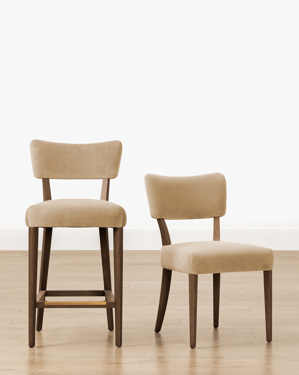 Two beige upholstered chairs with wooden legs sit on a light wood floor: the Makerspalm Etienne Stool (Ready to Ship), a tall, vintage-inspired kitchen stool, and a standard-height dining chair. Both feature curved backs and minimalist design from McGee & Co.