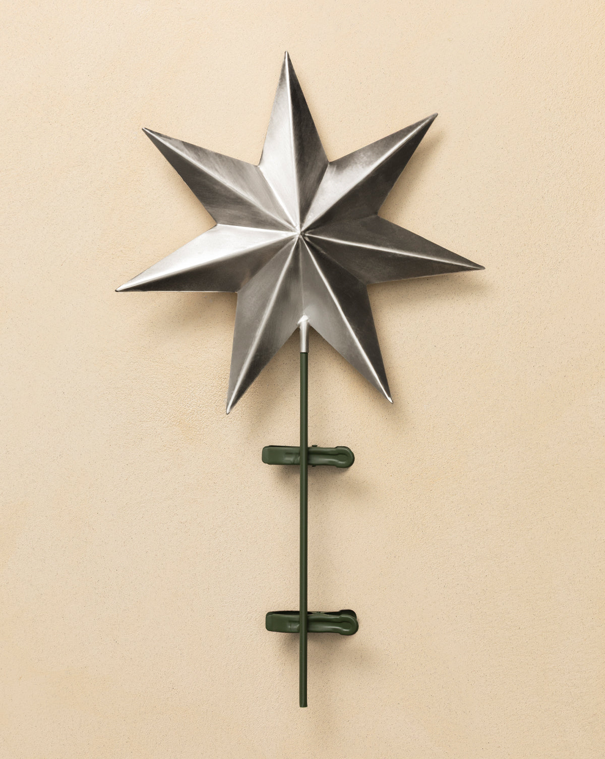 The Etoile Tree Topper features a metallic, eight-pointed design in an aged brass finish, mounted on a vertical green rod with two green clamps, displayed against a beige wall, by McGee & Co.