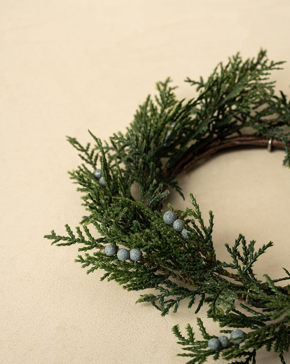 A partial view of the 8" Blueberry & Juniper Faux Wreath, featuring green pine branches and gray berries on a light beige background—an ideal faux greenery accent for elegant holiday decor, by McGee & Co.