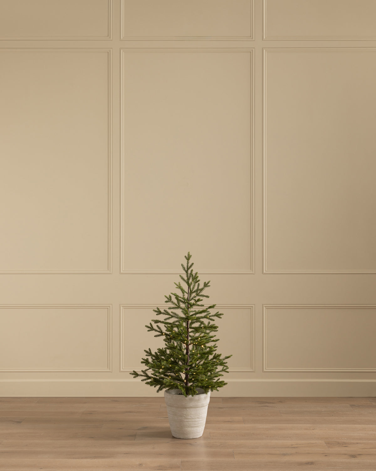 The Faux Spruce Pre-Lit Potted Tree is placed on a wooden floor before a beige paneled wall, serving as the centerpiece in a minimalist, empty room, by McGee & Co.