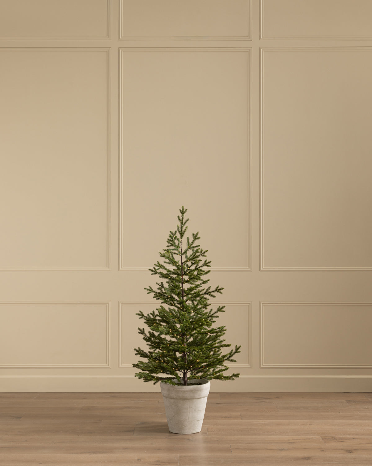 The Faux Spruce Pre-Lit Potted Tree stands on a wooden floor before a beige paneled wall, bringing greenery and soft light to the room, by McGee & Co.