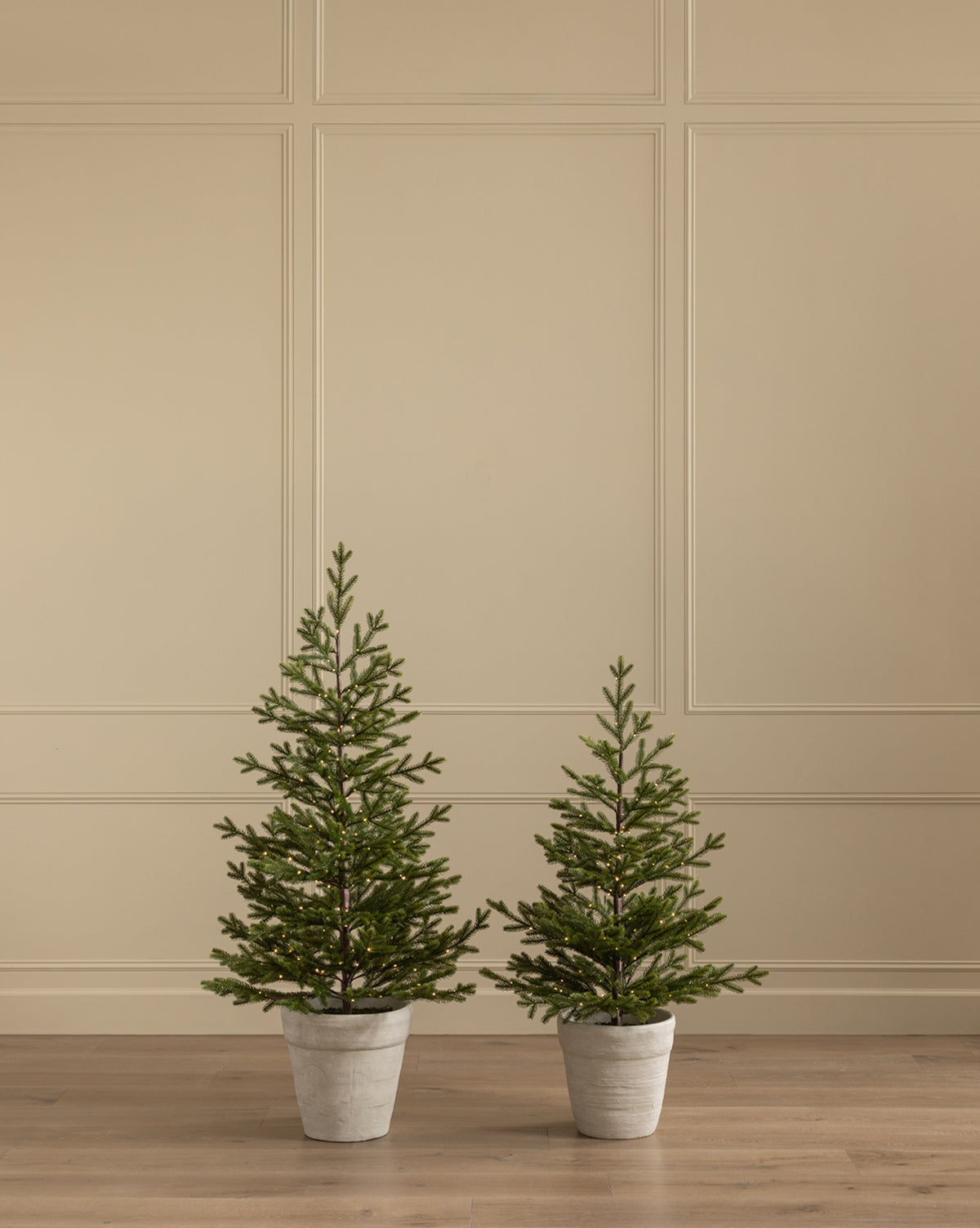 A Faux Spruce Pre-Lit Potted Tree and another small faux tree sit side by side on a wooden floor against a beige paneled wall, by McGee & Co.