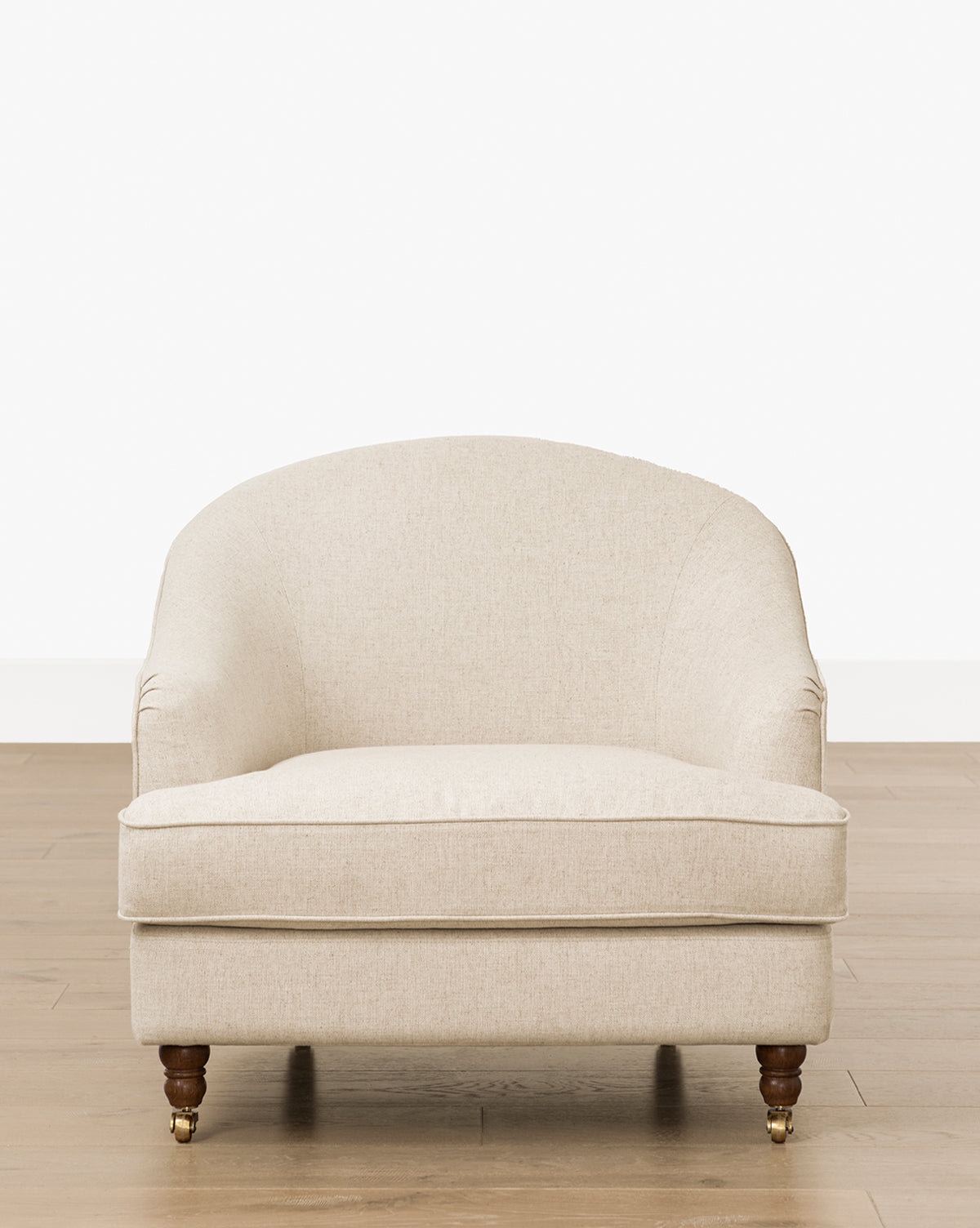 The Makerspalm Fletcher Lounge Chair (Ready to Ship) is a beige upholstered armchair with a rounded back, cushioned seat, and wooden legs with brass casters, shown on a light wood floor against a plain white background by McGee & Co.