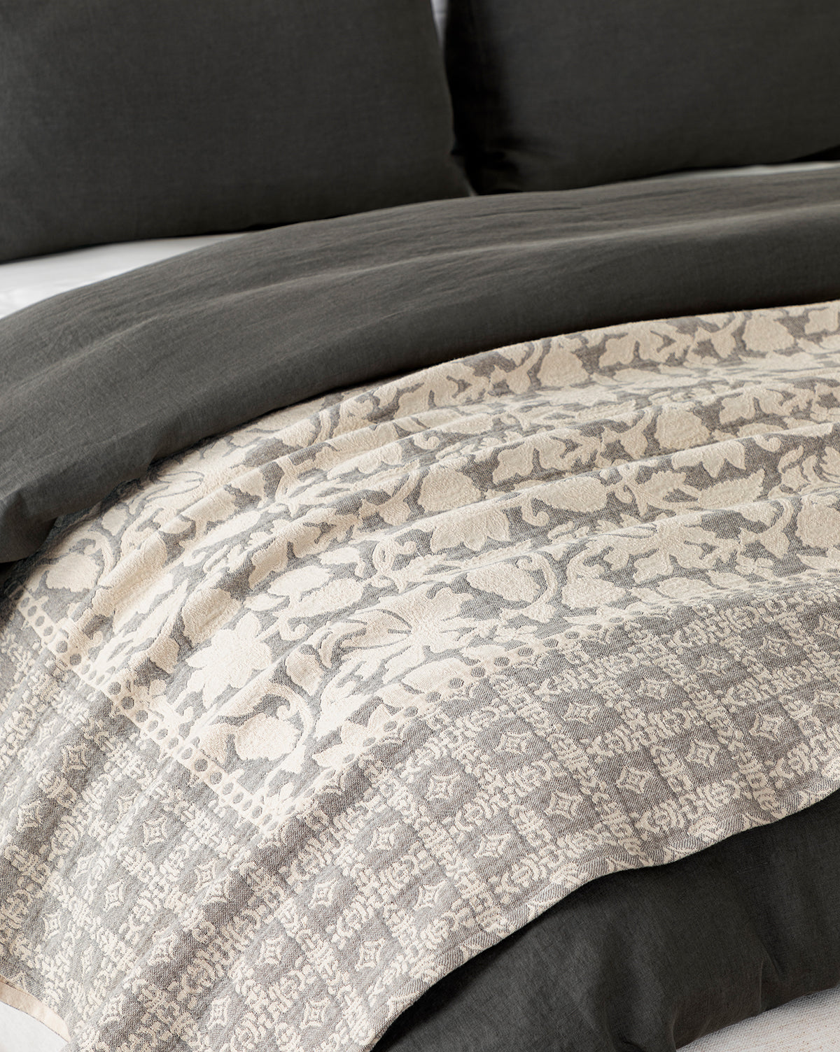 A close-up of a bed with dark gray pillows and the Floral Matelasse Coverlet, featuring a cream and light gray pattern with intricate floral and geometric designs for elegant bedding.