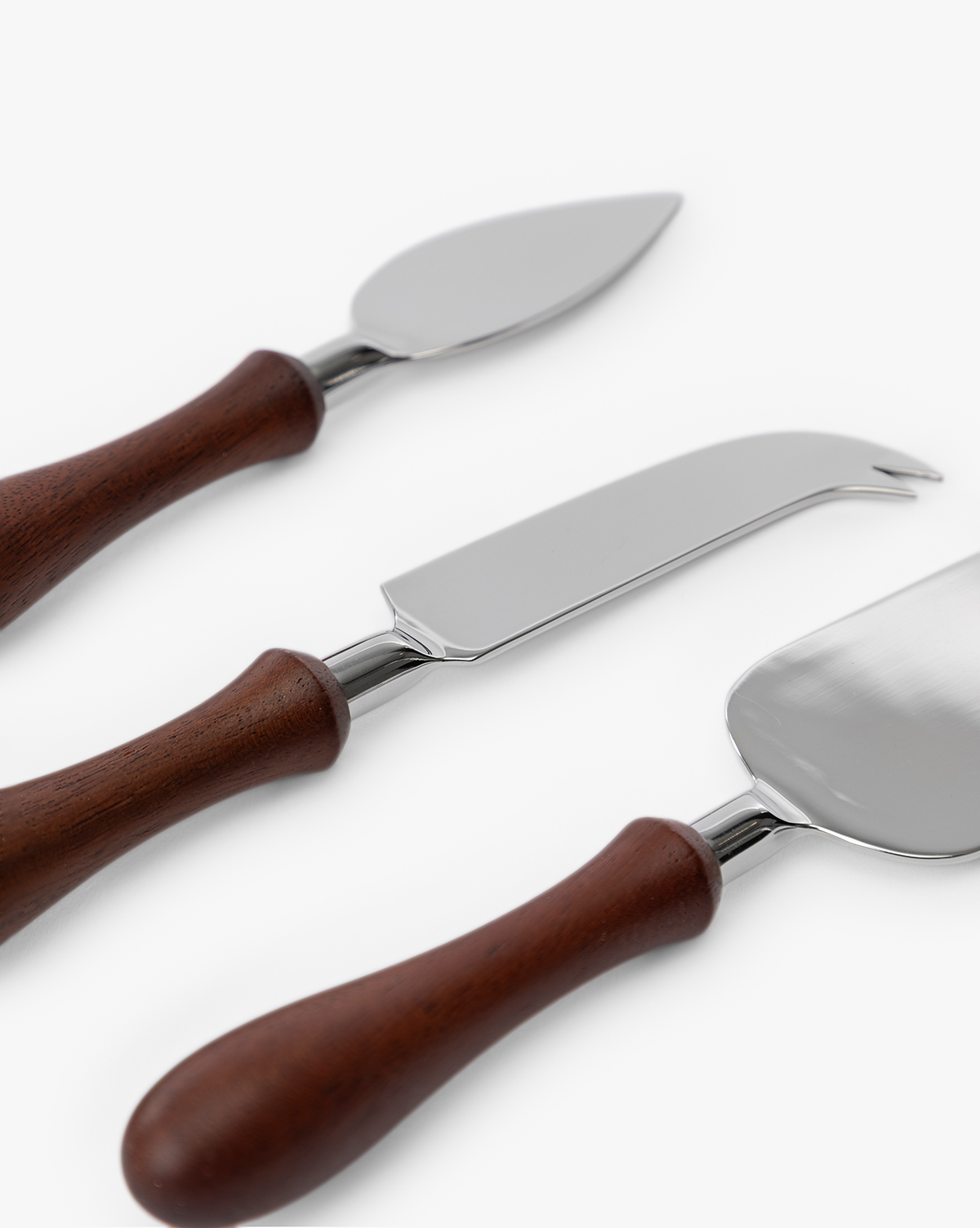 The Fromagerie Cheese Knives (Set of 3) feature stainless steel blades and wooden handles, each with a unique shape for slicing different cheeses. The three knives are arranged diagonally on a white background, by McGee & Co.