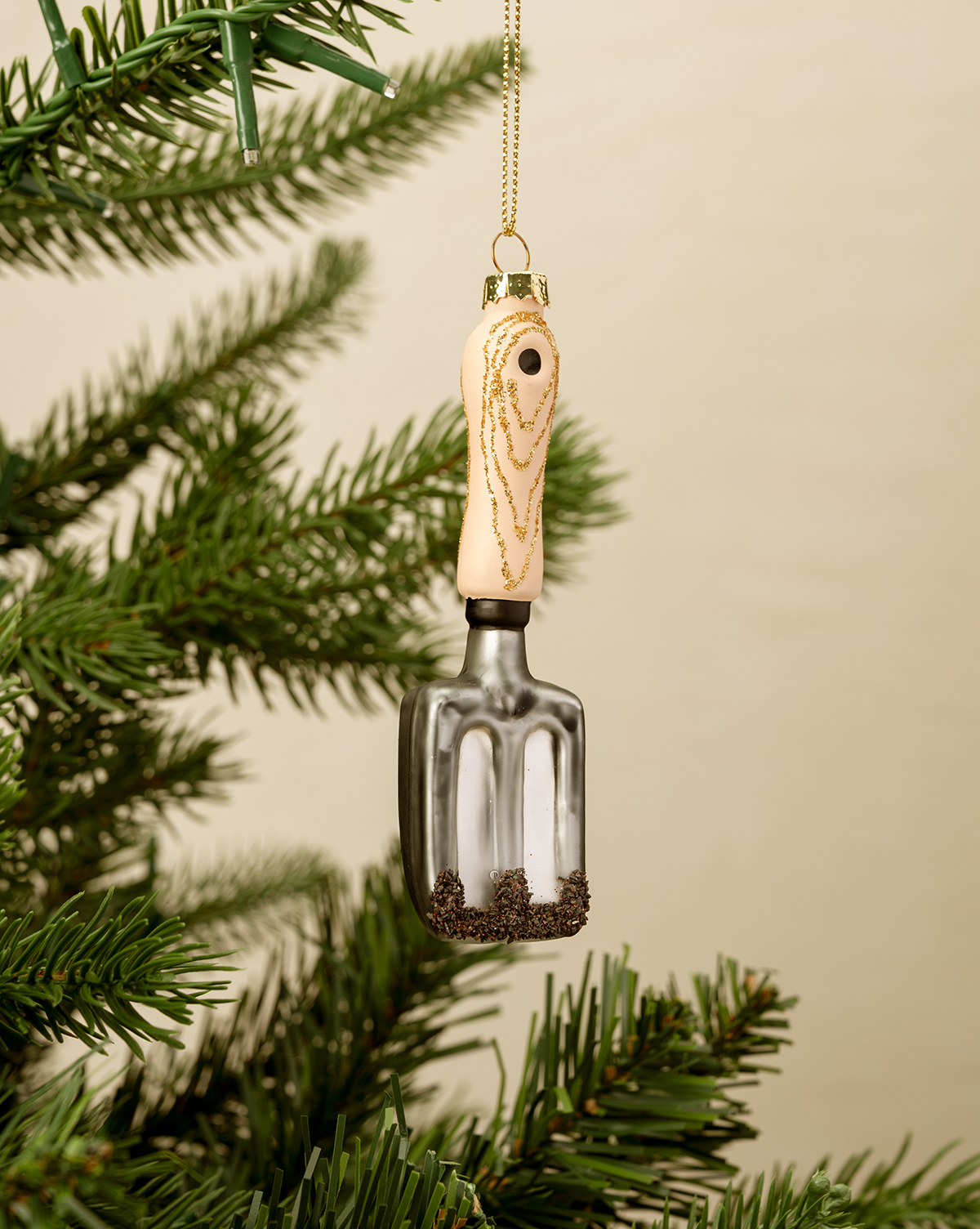 A Gardeners Ornament—a small trowel with a beige handle and metallic spade—hangs by a gold string on a Christmas tree, making it the perfect holiday gift for any gardener, by McGee & Co.