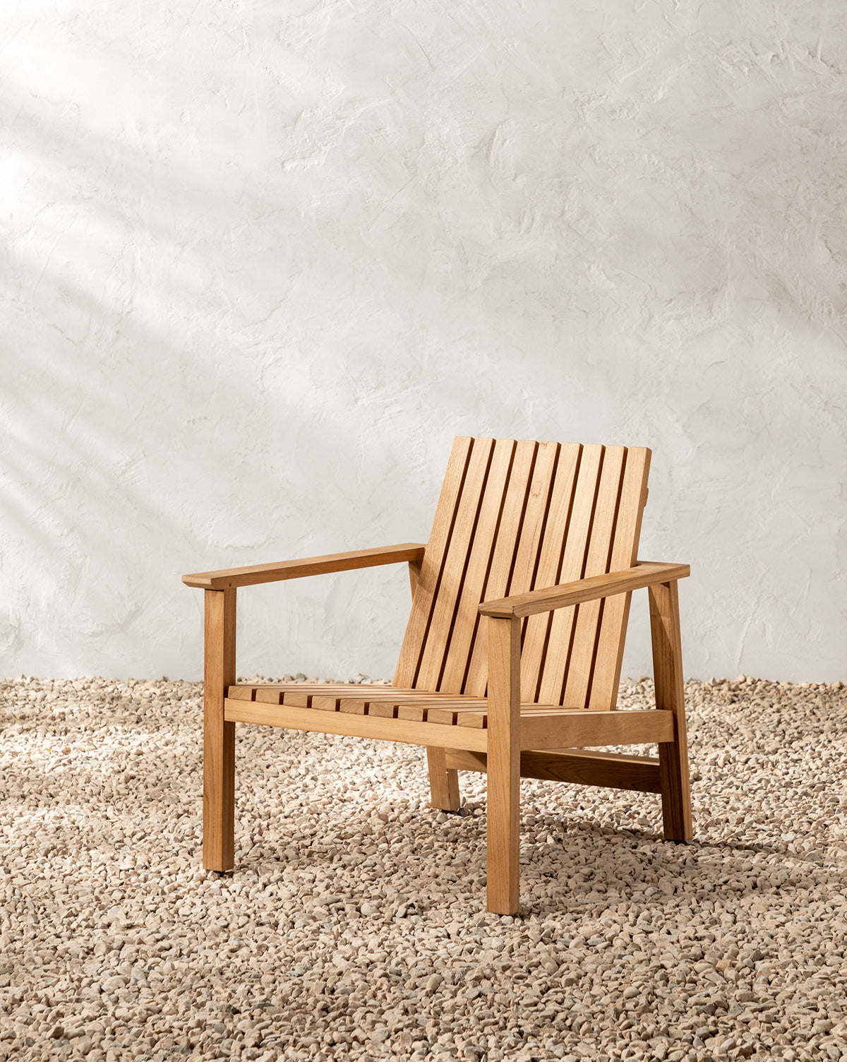 The Gilda Outdoor Lounge Chair, made of teak with a slatted back and seat, is set on gravel against a textured, light-toned wall in natural sunlight by McGee & Co.