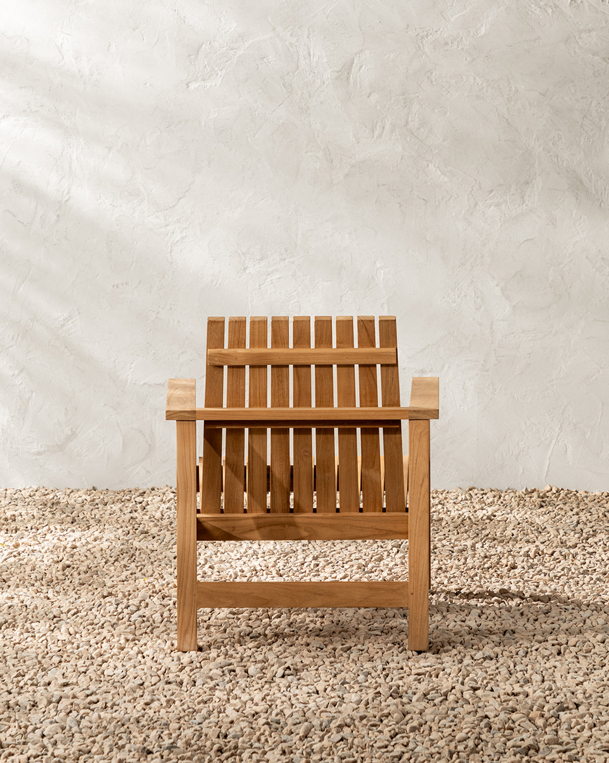 The Gilda Outdoor Lounge Chair, crafted from teak wood with vertical slats, sits on a gravel surface against a textured white wall by McGee & Co.