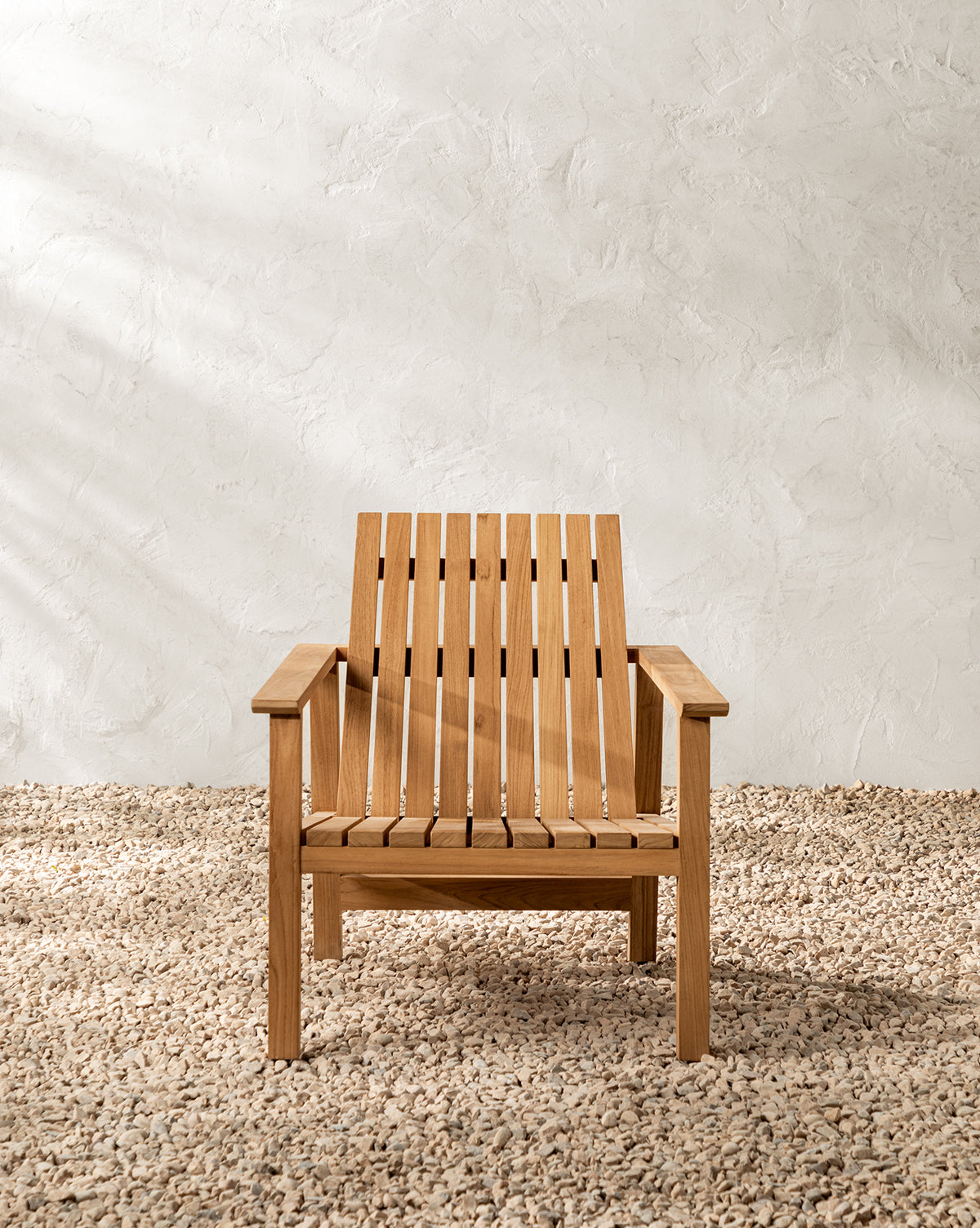 The Gilda Outdoor Lounge Chair, made of teak wood with a slatted seat and backrest, sits on gravel against a textured white wall in soft natural light, by McGee & Co.