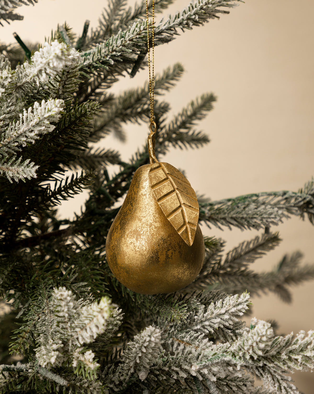 The Gilded Pear Ornament, featuring a detailed leaf, hangs from a snow-dusted evergreen branch against a neutral background, by McGee & Co.