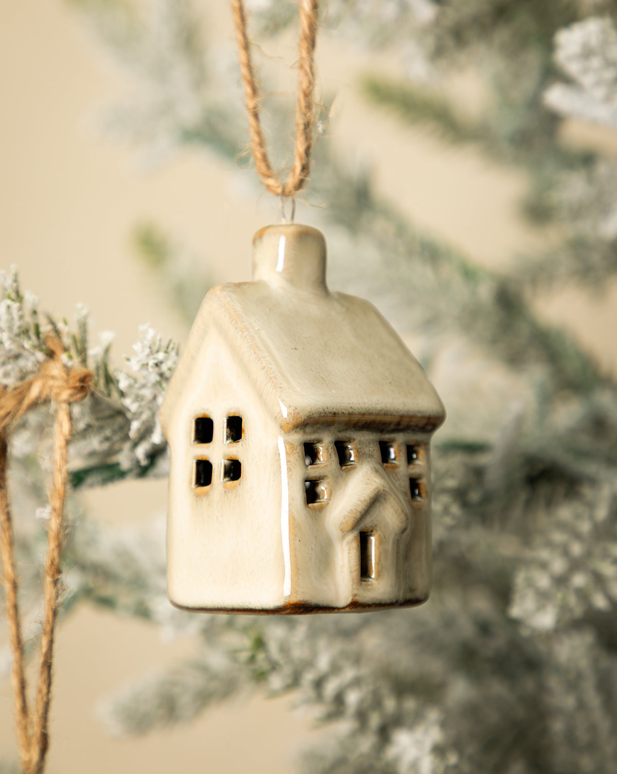 The Glazed Porcelain Houses (Set of 2) hang from twine on a snow-dusted Christmas tree branch, featuring doors and windows for a charming, festive touch to your holiday décor, by McGee & Co.