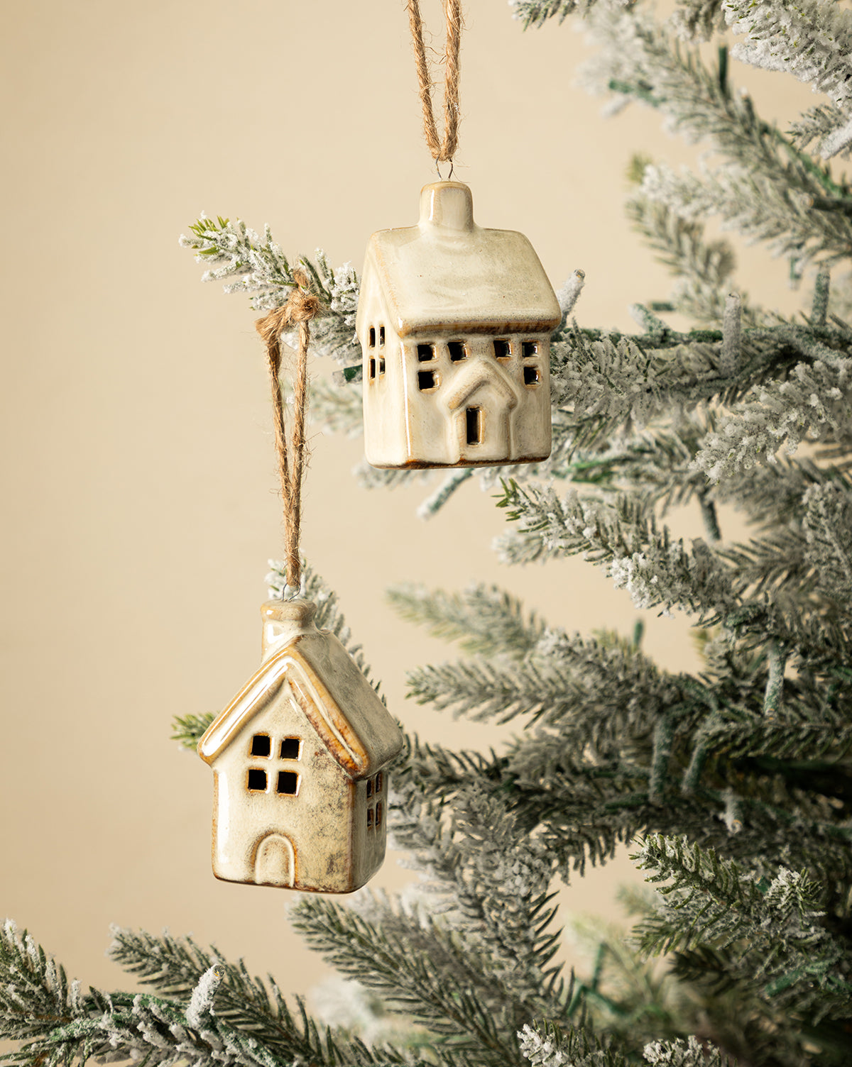 The Glazed Porcelain Houses (Set of 2) hang from twine on a snow-dusted Christmas tree branch against a beige background, bringing charming festive decor to your holiday setting, by McGee & Co.