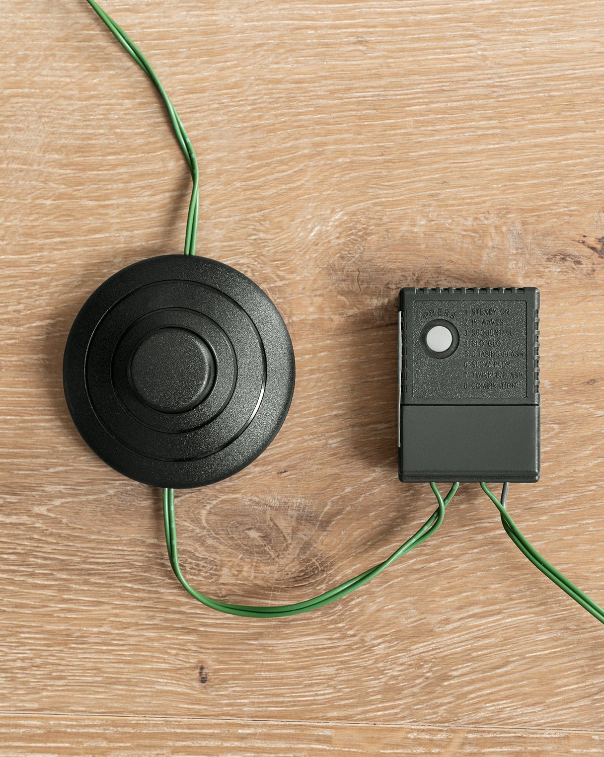 A black round foot pedal and a small rectangular control box, connected by green wires, are used to operate the 9' Grandis Fir Pre-Lit Faux Tree and are shown on a light wooden surface, by McGee & Co.