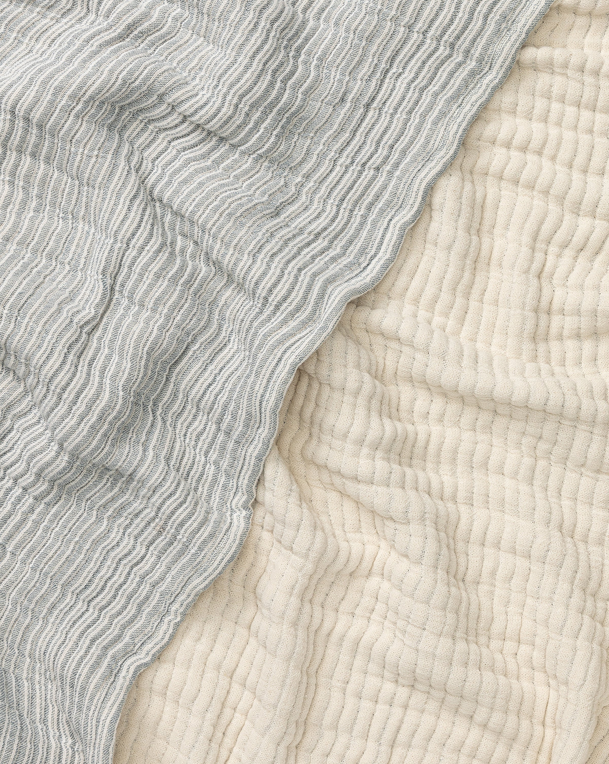 A close-up of Hanson Striped Cotton Coverlet beside an off-white quilted fabric showcases the contrast between its light gray wavy stripes and the crinkled, textured look of neutral bedding styles.