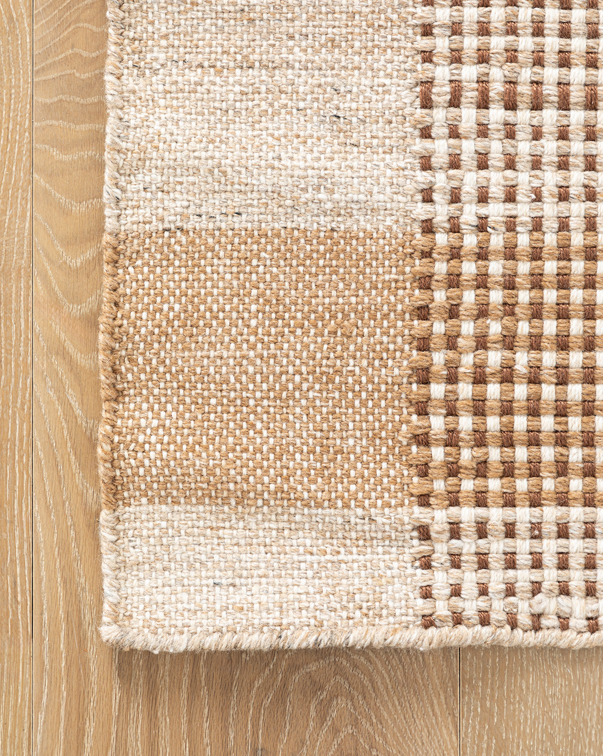 A close-up of the Hargrove Handwoven Indoor/Outdoor Rug, showcasing its beige, brown, and cream geometric checker pattern with solid and checkered sections for added texture, displayed on a light wood floor.